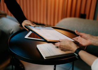 Zwei Personen sitzen an einem runden schwarzen Tisch. Eine Person überreicht der anderen ein Klemmbrett mit Dokumenten. Auf dem Tisch liegen außerdem ein kariertes Notizbuch und ein teilweise verdecktes Tablet oder Laptop. Im Hintergrund sind gepolsterte Möbel und vertikale Holzlamellen an der Wand zu sehen.