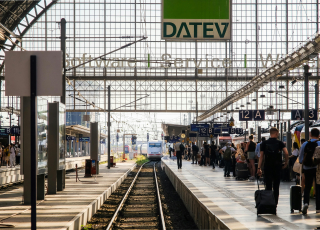 Ein belebter Bahnhof mit mehreren Gleisen und Bahnsteigen. Menschen bewegen sich durch die Szene, einige warten, andere tragen Gepäck. Im Hintergrund fährt ein Zug in den Bahnhof ein. Über den Bahnsteigen befindet sich ein großes Glasdach, das Tageslicht hereinlässt. Ein auffälliges grünes Schild mit der Aufschrift ‚DATEV‘ hängt über dem Bereich. Plattformschilder wie ‚12 A‘ und ‚12 B‘ sind ebenfalls sichtbar.