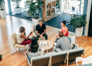Fünf Personen sitzen in einer modernen Lounge und unterhalten sich an einem runden Tisch, umgeben von Pflanzen, Regalen und großen Fenstern mit Tageslicht.
