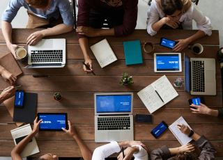 Eine Gruppe arbeitet gemeinsam mit Laptops, Tablets und Notizblöcken an einem Tisch, digitale Zusammenarbeit steht im Fokus.