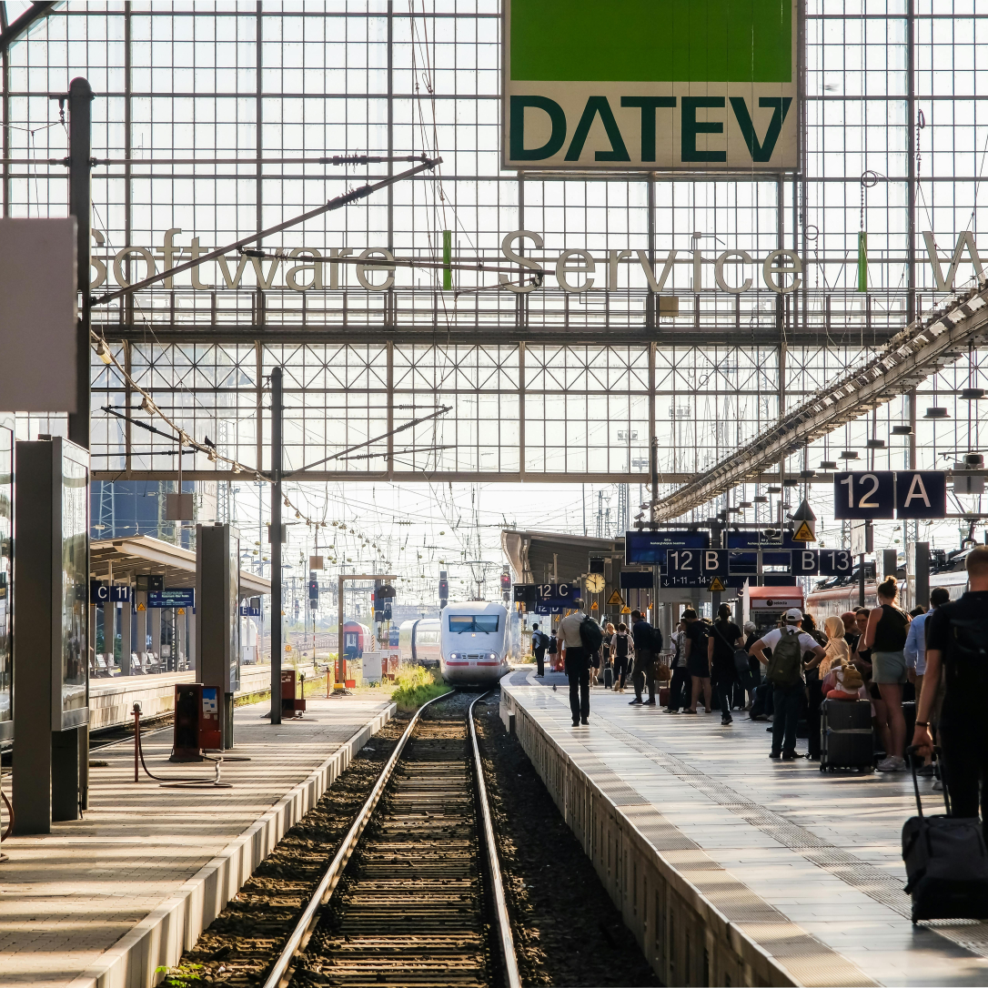 Ein belebter Bahnhof mit mehreren Gleisen und Bahnsteigen. Menschen bewegen sich durch die Szene, einige warten, andere tragen Gepäck. Im Hintergrund fährt ein Zug in den Bahnhof ein. Über den Bahnsteigen befindet sich ein großes Glasdach, das Tageslicht hereinlässt. Ein auffälliges grünes Schild mit der Aufschrift ‚DATEV‘ hängt über dem Bereich. Plattformschilder wie ‚12 A‘ und ‚12 B‘ sind ebenfalls sichtbar.