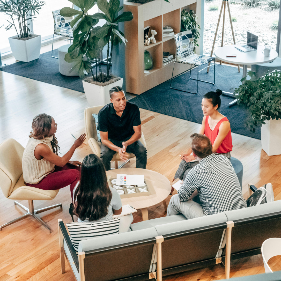 Fünf Personen sitzen in einer modernen Lounge und unterhalten sich an einem runden Tisch, umgeben von Pflanzen, Regalen und großen Fenstern mit Tageslicht.